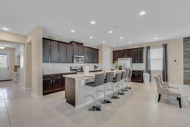 kitchen with appliances with stainless steel finishes, light tile patterned floors, a kitchen breakfast bar, dark brown cabinets, and a center island with sink