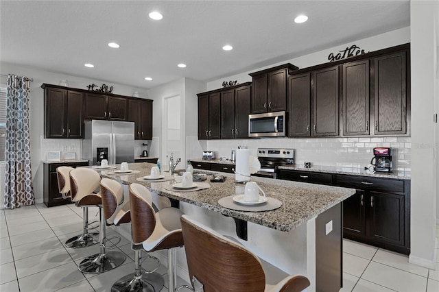 kitchen featuring appliances with stainless steel finishes, light tile patterned floors, a kitchen breakfast bar, backsplash, and an island with sink