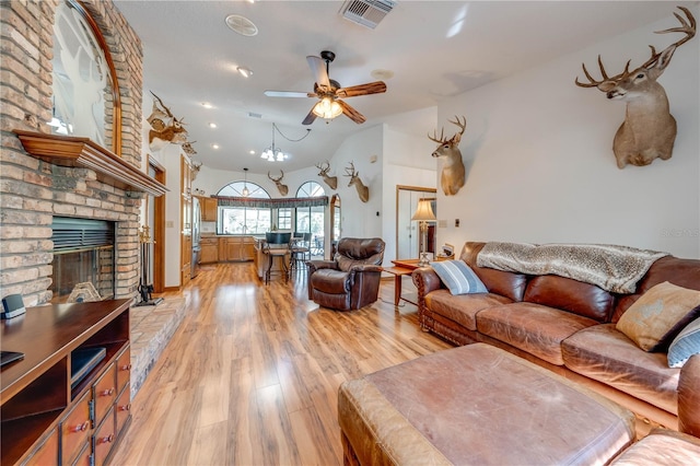 living room with ceiling fan with notable chandelier, lofted ceiling, light wood-type flooring, and a brick fireplace