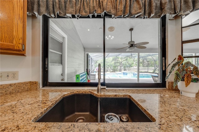 kitchen featuring brown cabinets, a sunroom, ceiling fan, a sink, and light stone countertops