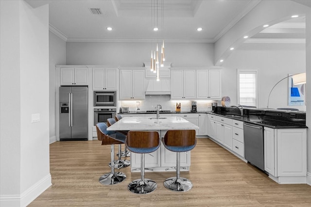 kitchen featuring dark stone countertops, ornamental molding, pendant lighting, stainless steel appliances, and a kitchen island with sink