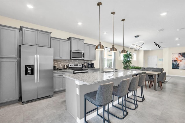 kitchen with sink, gray cabinetry, stainless steel appliances, light stone counters, and an island with sink