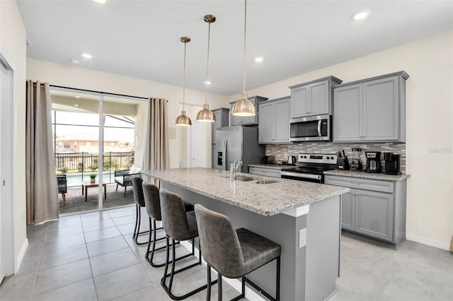 kitchen featuring sink, hanging light fixtures, stainless steel appliances, light stone counters, and an island with sink