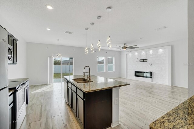 kitchen with stainless steel appliances, a center island with sink, light stone counters, light wood-type flooring, and decorative light fixtures