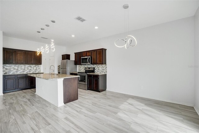 kitchen with stainless steel appliances, light stone countertops, an island with sink, pendant lighting, and decorative backsplash