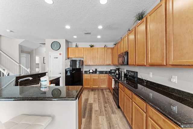 kitchen with light hardwood / wood-style floors, dark stone counters, black appliances, and a kitchen island with sink