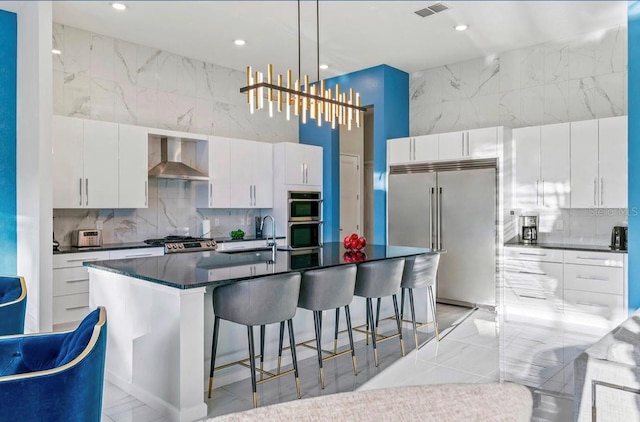 kitchen featuring wall chimney exhaust hood, white cabinetry, stainless steel appliances, an island with sink, and sink