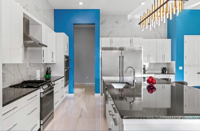 kitchen with appliances with stainless steel finishes, wall chimney exhaust hood, sink, decorative light fixtures, and white cabinetry