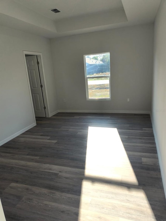 empty room with dark wood-type flooring, a tray ceiling, visible vents, and baseboards