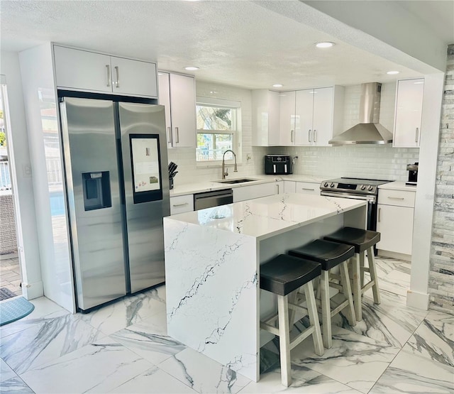 kitchen featuring a breakfast bar area, appliances with stainless steel finishes, white cabinetry, a kitchen island, and wall chimney exhaust hood