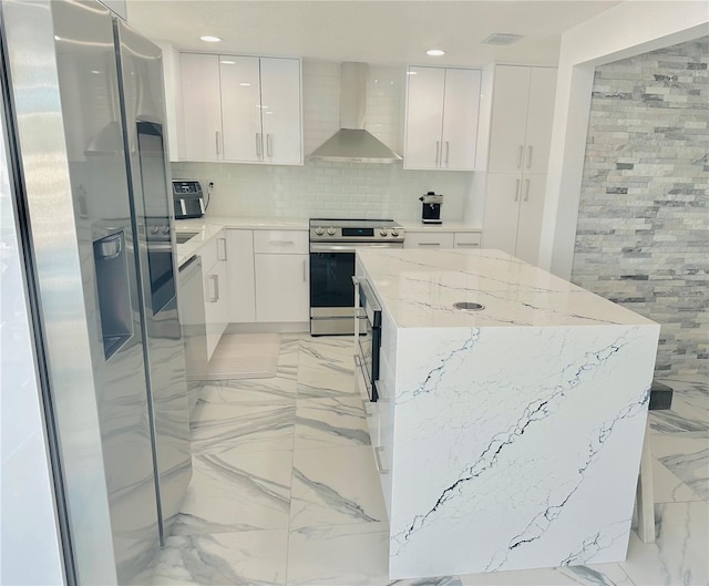 kitchen featuring wall chimney range hood, appliances with stainless steel finishes, white cabinetry, light stone countertops, and a kitchen island