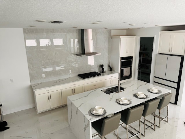 kitchen with stainless steel gas cooktop, light stone counters, a center island with sink, and wall chimney exhaust hood