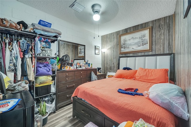 bedroom with a textured ceiling, ceiling fan, wooden walls, and light hardwood / wood-style floors