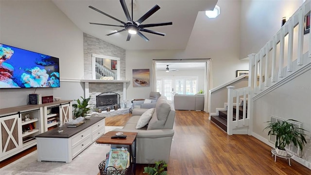 living room featuring a ceiling fan, wood finished floors, stairs, a stone fireplace, and high vaulted ceiling