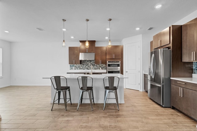 kitchen featuring pendant lighting, backsplash, stainless steel appliances, dark brown cabinetry, and an island with sink