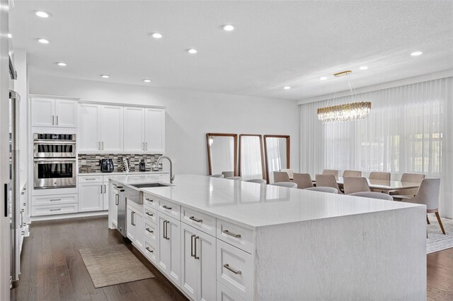 kitchen featuring dark wood-type flooring, stainless steel double oven, hanging light fixtures, white cabinetry, and a center island with sink