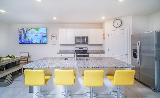 kitchen with light tile patterned flooring, stainless steel appliances, a kitchen island with sink, white cabinetry, and light stone counters