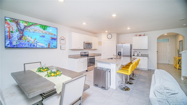 kitchen with white cabinetry, appliances with stainless steel finishes, a kitchen island with sink, light stone countertops, and light tile patterned floors