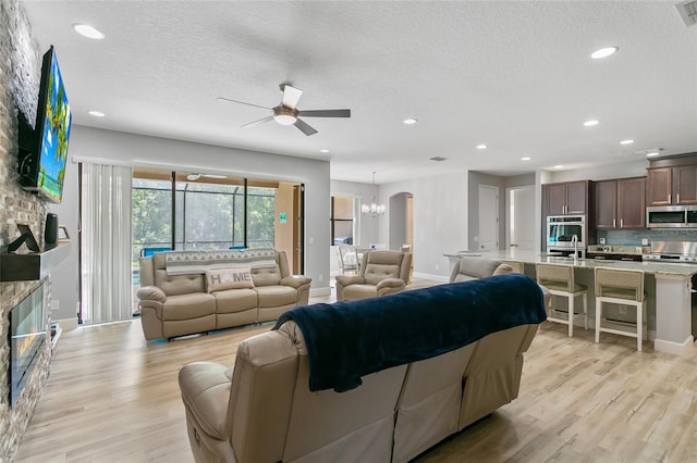 living room featuring ceiling fan with notable chandelier, a stone fireplace, light hardwood / wood-style flooring, and a textured ceiling