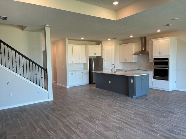 kitchen with white cabinetry, a center island with sink, refrigerator, wall chimney exhaust hood, and double oven