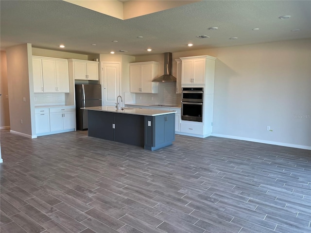 kitchen with an island with sink, white cabinetry, wall chimney exhaust hood, double oven, and black fridge