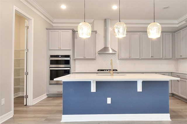 kitchen with light wood-type flooring, stainless steel double oven, wall chimney exhaust hood, and an island with sink