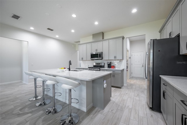 kitchen with gray cabinetry, light stone countertops, a center island with sink, appliances with stainless steel finishes, and light hardwood / wood-style floors