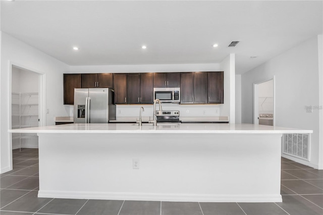 kitchen with a kitchen island with sink, dark tile patterned flooring, dark brown cabinets, and appliances with stainless steel finishes