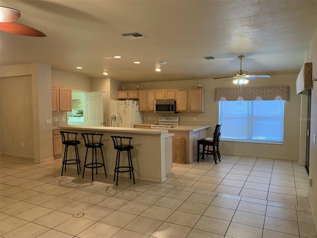 kitchen with light brown cabinetry, light tile patterned floors, white appliances, ceiling fan, and a breakfast bar