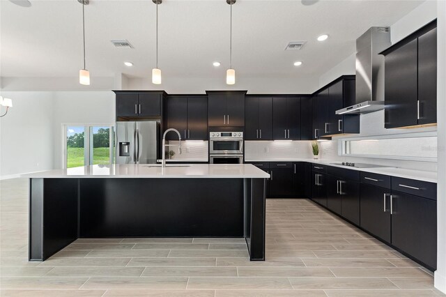 kitchen featuring pendant lighting, stainless steel appliances, wall chimney exhaust hood, and a center island with sink