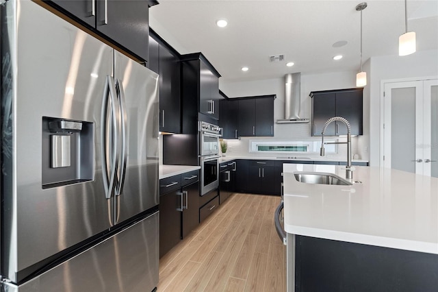 kitchen featuring sink, a kitchen island with sink, hanging light fixtures, stainless steel appliances, and wall chimney exhaust hood