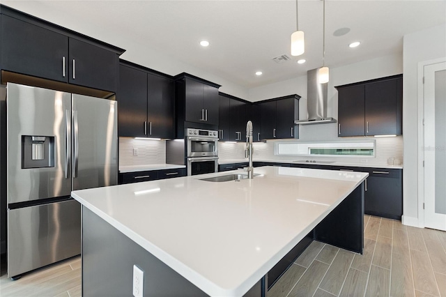 kitchen with sink, a center island with sink, pendant lighting, stainless steel appliances, and wall chimney range hood