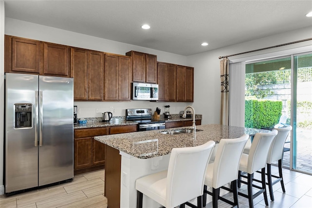 kitchen with a kitchen bar, light stone counters, an island with sink, sink, and appliances with stainless steel finishes