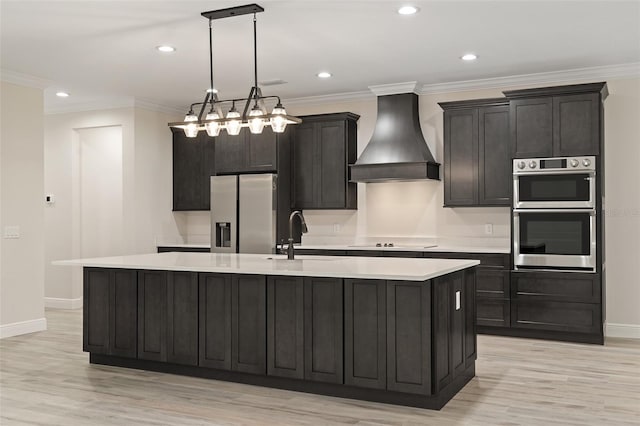 kitchen with appliances with stainless steel finishes, custom range hood, hanging light fixtures, an island with sink, and light wood-type flooring