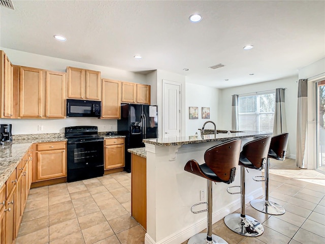 kitchen with a center island with sink, black appliances, light tile patterned floors, a kitchen bar, and light stone counters