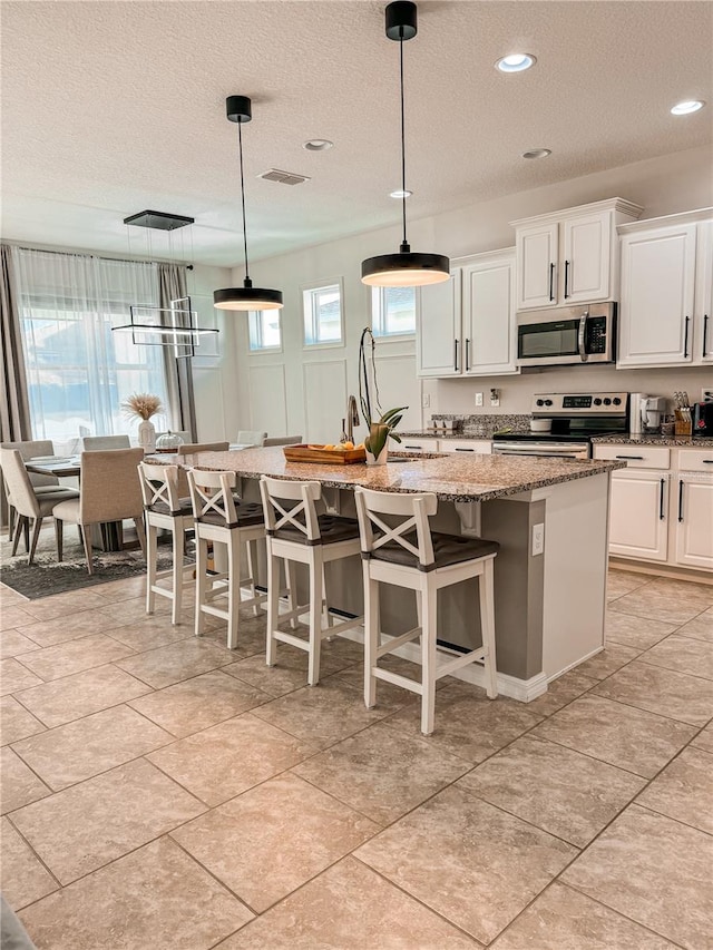 kitchen with white cabinets, hanging light fixtures, light stone counters, stainless steel appliances, and a center island with sink