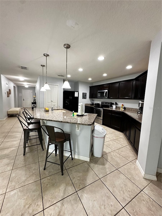 kitchen with sink, hanging light fixtures, stainless steel appliances, a center island with sink, and light stone countertops