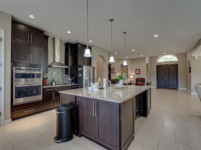 kitchen featuring dark brown cabinets, pendant lighting, wall chimney exhaust hood, a center island with sink, and appliances with stainless steel finishes