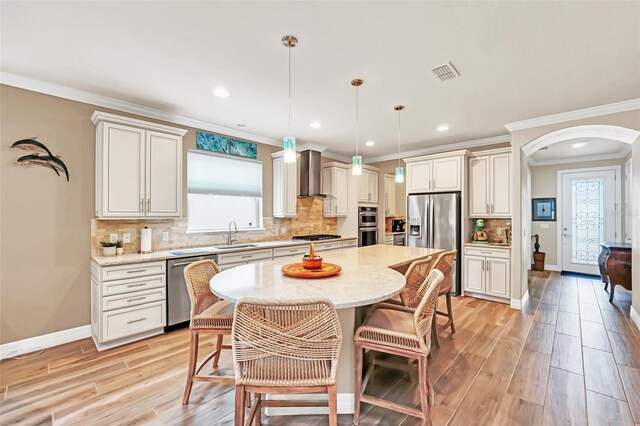 kitchen with light hardwood / wood-style floors, a kitchen island, wall chimney range hood, stainless steel appliances, and decorative light fixtures