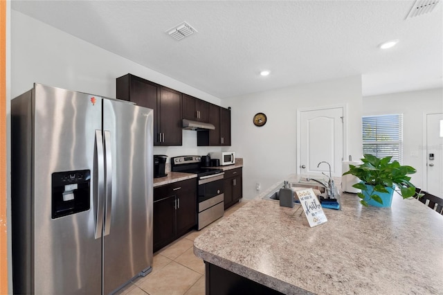 kitchen with dark brown cabinets, a textured ceiling, an island with sink, stainless steel appliances, and light tile patterned floors