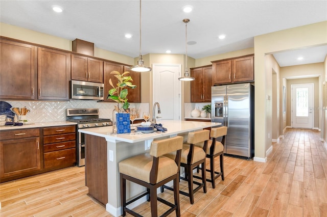 kitchen with decorative light fixtures, a kitchen island with sink, stainless steel appliances, light hardwood / wood-style floors, and decorative backsplash