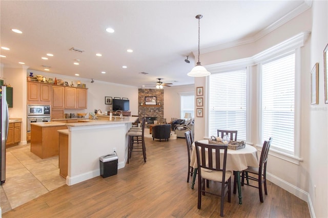 dining space featuring ceiling fan, a stone fireplace, light wood-type flooring, and ornamental molding