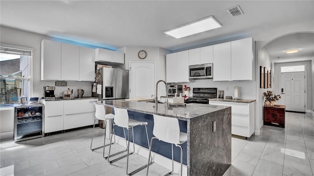 kitchen with appliances with stainless steel finishes, a kitchen island with sink, wine cooler, and white cabinetry