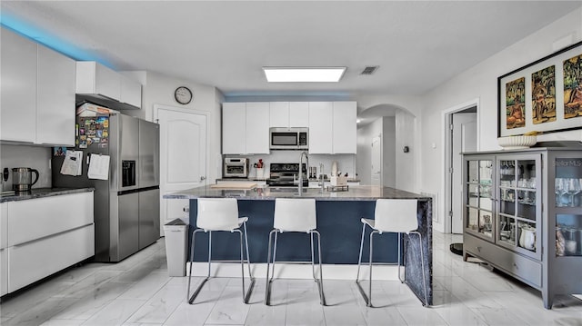 kitchen featuring an island with sink, white cabinets, appliances with stainless steel finishes, and dark stone counters