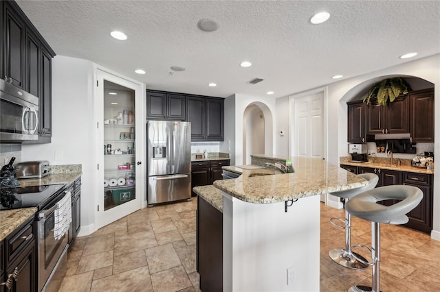 kitchen featuring light stone counters, a breakfast bar, stainless steel appliances, a textured ceiling, and a center island with sink