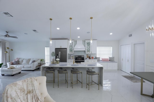 kitchen featuring pendant lighting, a kitchen island with sink, stainless steel appliances, custom range hood, and white cabinets