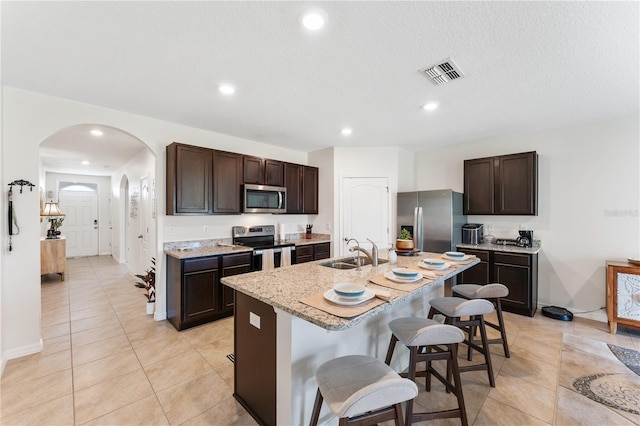 kitchen with sink, dark brown cabinets, a kitchen island with sink, appliances with stainless steel finishes, and a kitchen bar