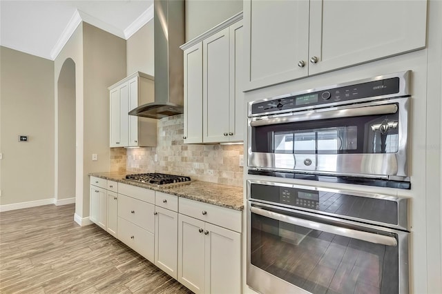 kitchen featuring light stone countertops, stainless steel appliances, wall chimney range hood, light hardwood / wood-style flooring, and white cabinetry