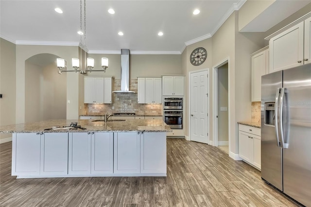 kitchen featuring hanging light fixtures, stainless steel appliances, wall chimney range hood, an island with sink, and light hardwood / wood-style floors