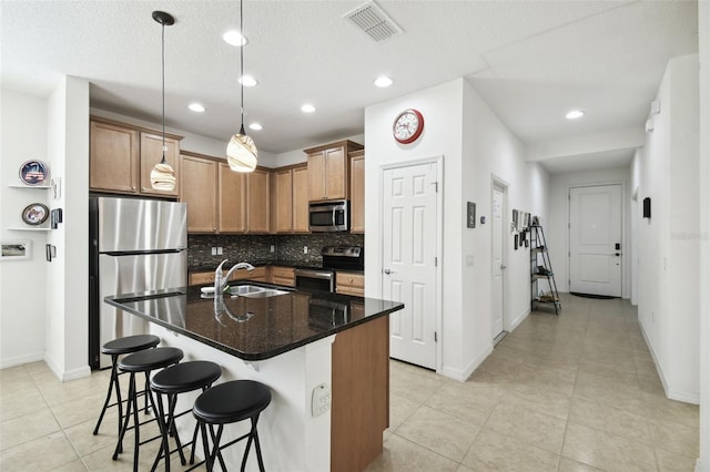 kitchen with a textured ceiling, a kitchen island with sink, sink, stainless steel appliances, and dark stone countertops